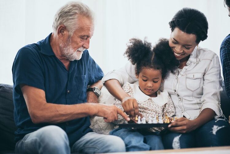 grandfather sitting on the couch with his granddaughter and daughter in law, playing chess - lovely multi-racial family - loneliness