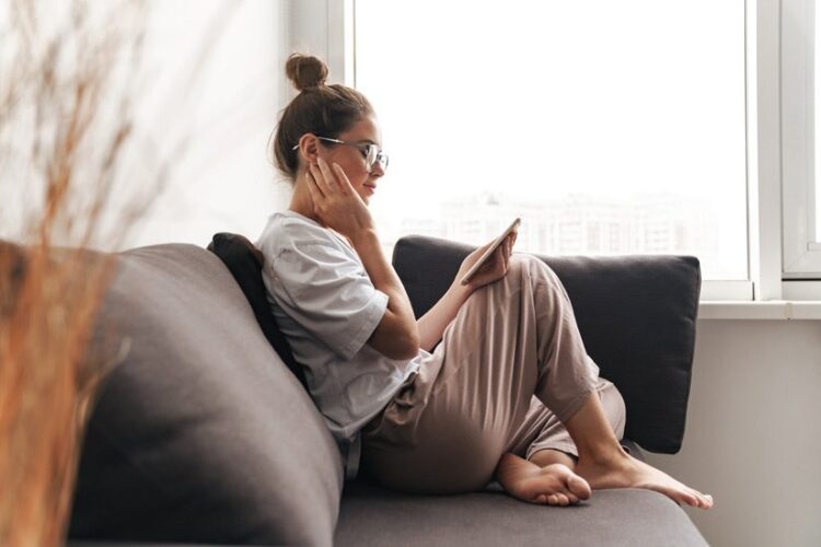 pretty young woman sitting on the couch at home looking at her smart phone, relaxing alone - newly sober