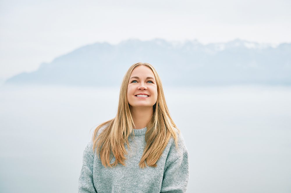 Outdoor portrait of happy beautiful young woman relaxing in mountains over the clouds, wearing grey pullover