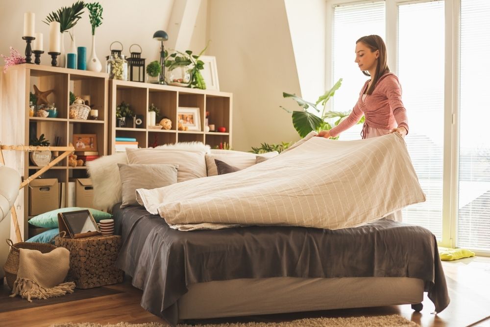 Woman making a bed in a bright, organized bedroom filled with plants, books, and home decor.