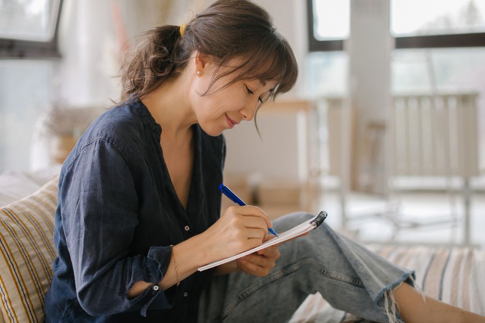Woman sitting comfortably indoors, smiling as she writes in a notebook