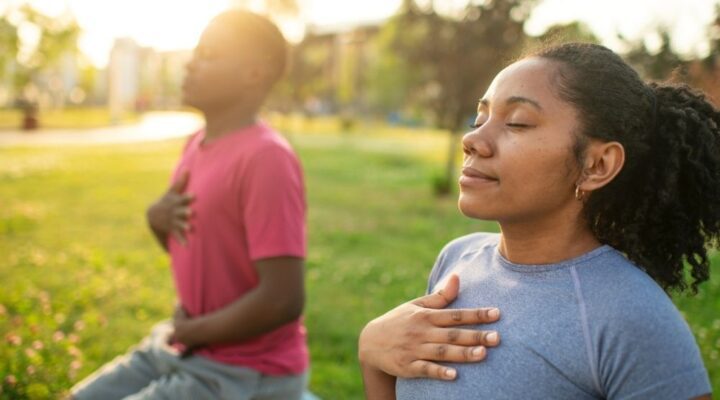 Two people sit outdoors on yoga mats with eyes closed, each placing one hand on their chest while practicing mindful breathing in a sunlit park.