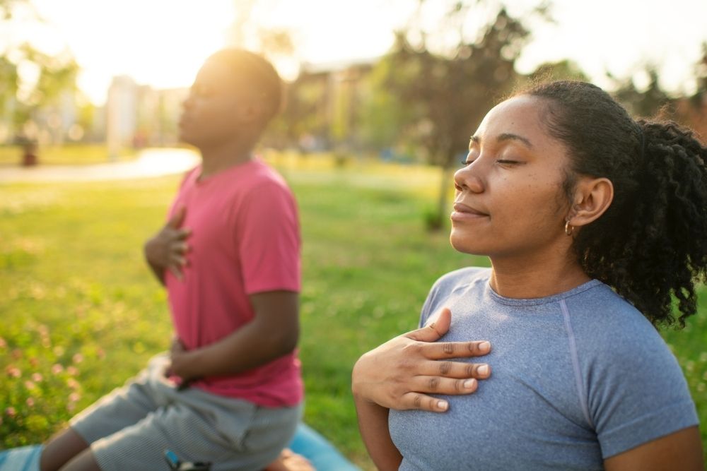Two people sit outdoors on yoga mats with eyes closed, each placing one hand on their chest while practicing mindful breathing in a sunlit park.