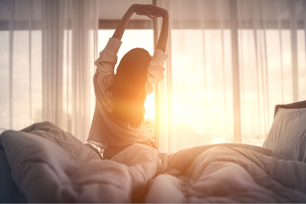 Woman stretching in bed at sunrise, with soft morning light filtering through sheer curtains, creating a calm and peaceful atmosphere.