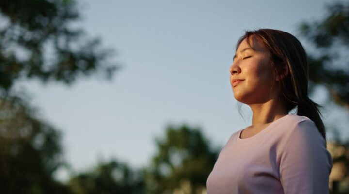A woman stands outdoors with her eyes closed and a peaceful expression, enjoying the fresh air and sunlight. Trees and blue sky are in the background.