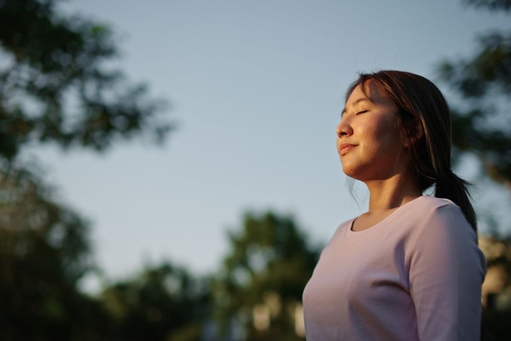 A woman stands outdoors with her eyes closed and a peaceful expression, enjoying the fresh air and sunlight. Trees and blue sky are in the background.