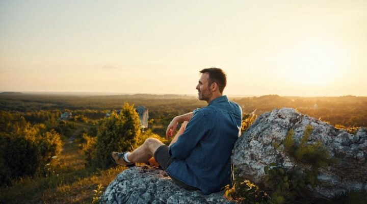 Man sitting on a rocky hilltop at sunset, looking out over the landscape.