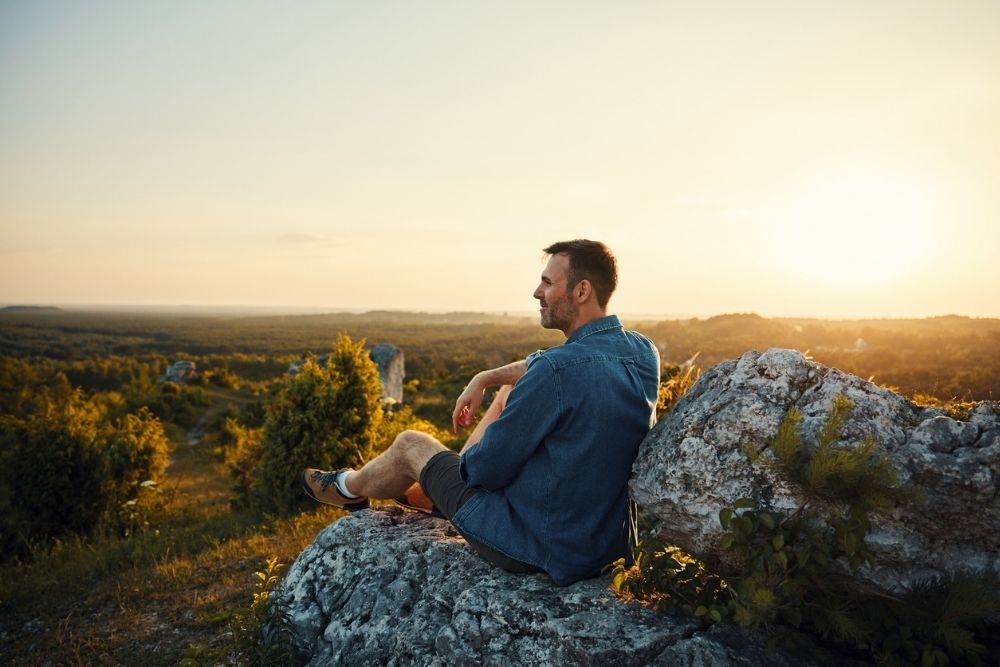 Man sitting on a rocky hilltop at sunset, looking out over the landscape.