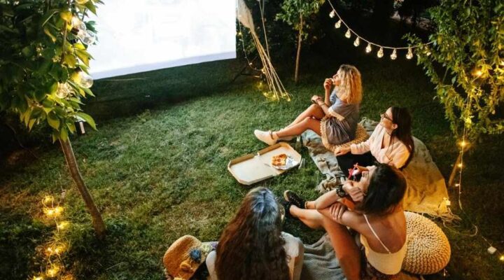 A group of friends sitting on blankets in a backyard at night watching an outdoor movie projected on a screen, surrounded by string lights and trees.