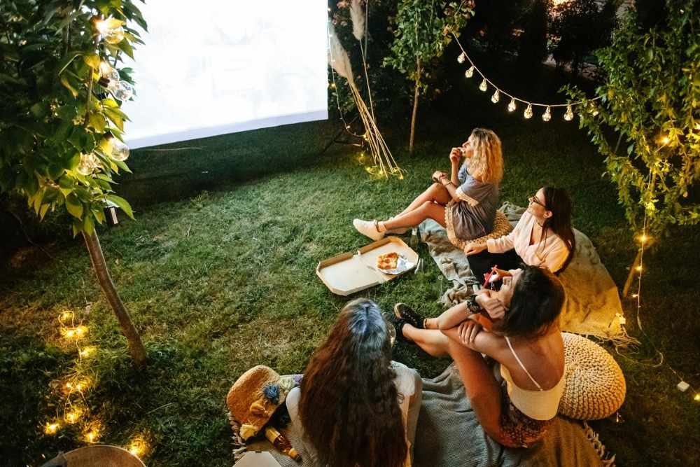 A group of friends sitting on blankets in a backyard at night watching an outdoor movie projected on a screen, surrounded by string lights and trees.