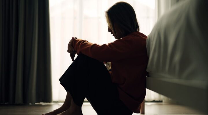 Silhouette of a woman sitting on the floor by a bed, with her knees drawn up and head resting on her arms, appearing sad or reflective.