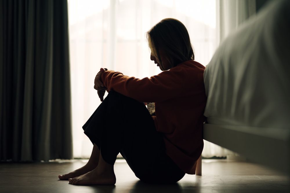 Silhouette of a woman sitting on the floor by a bed, with her knees drawn up and head resting on her arms, appearing sad or reflective.