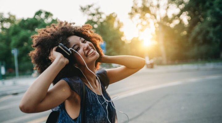 A joyful young woman with curly hair listens to music on headphones outdoors at sunset, smiling and holding her phone.