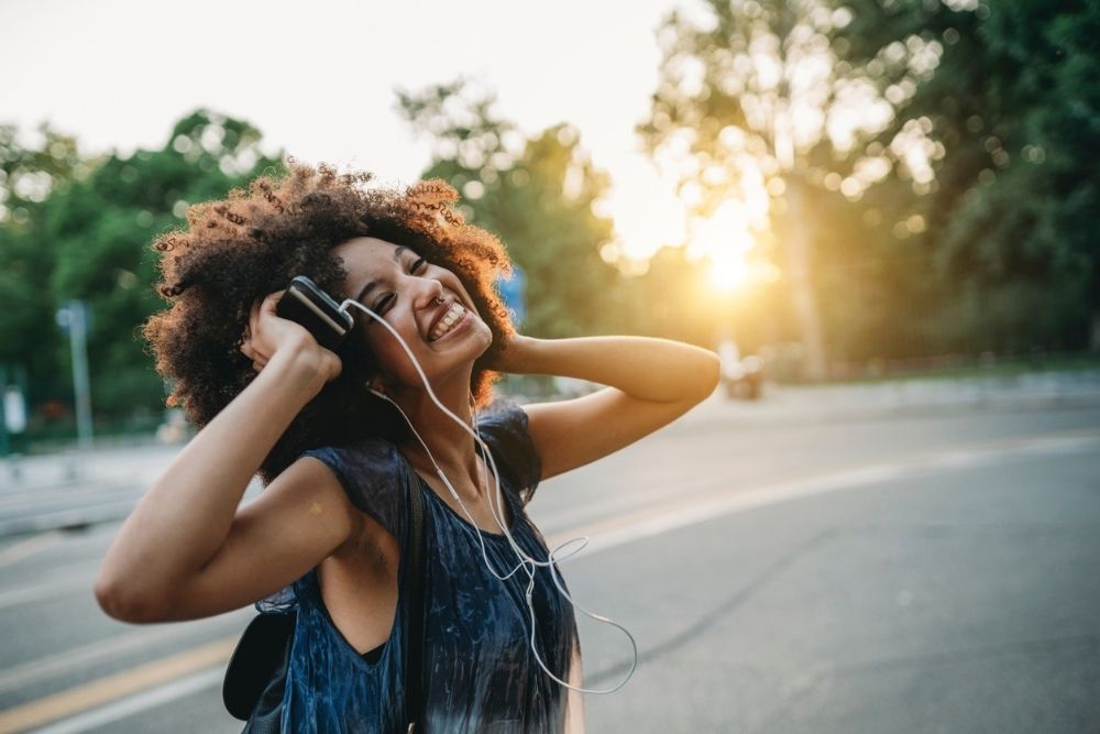 A joyful young woman with curly hair listens to music on headphones outdoors at sunset, smiling and holding her phone.