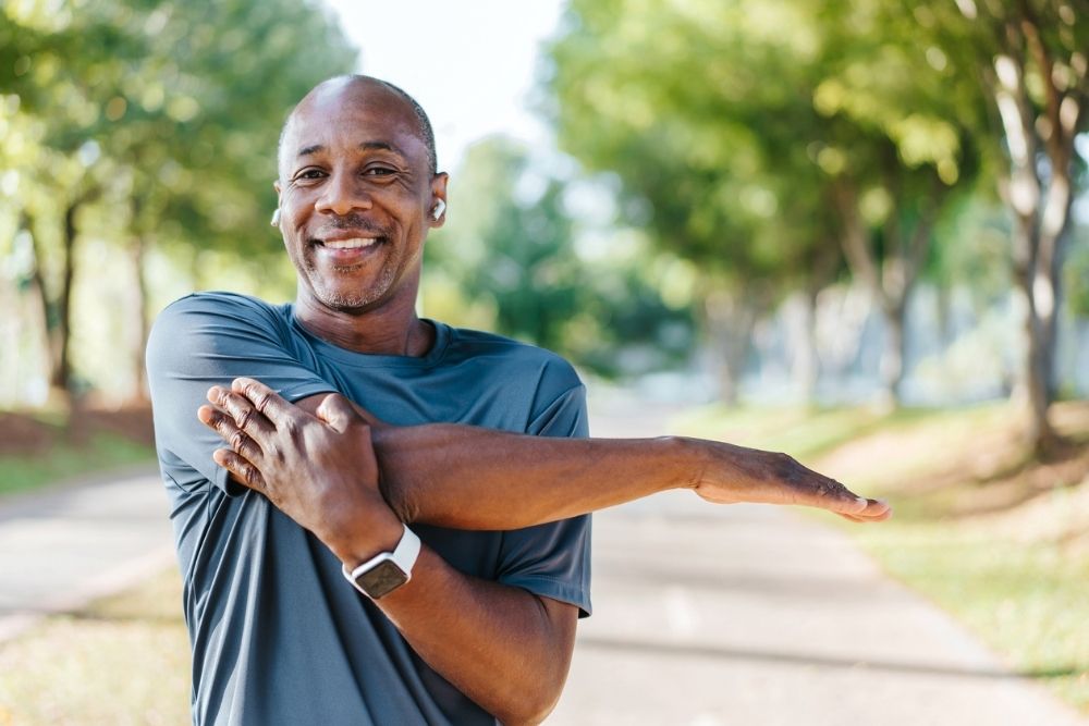 Smiling man in athletic wear stretching his arm outdoors on a sunny day, preparing for exercise.