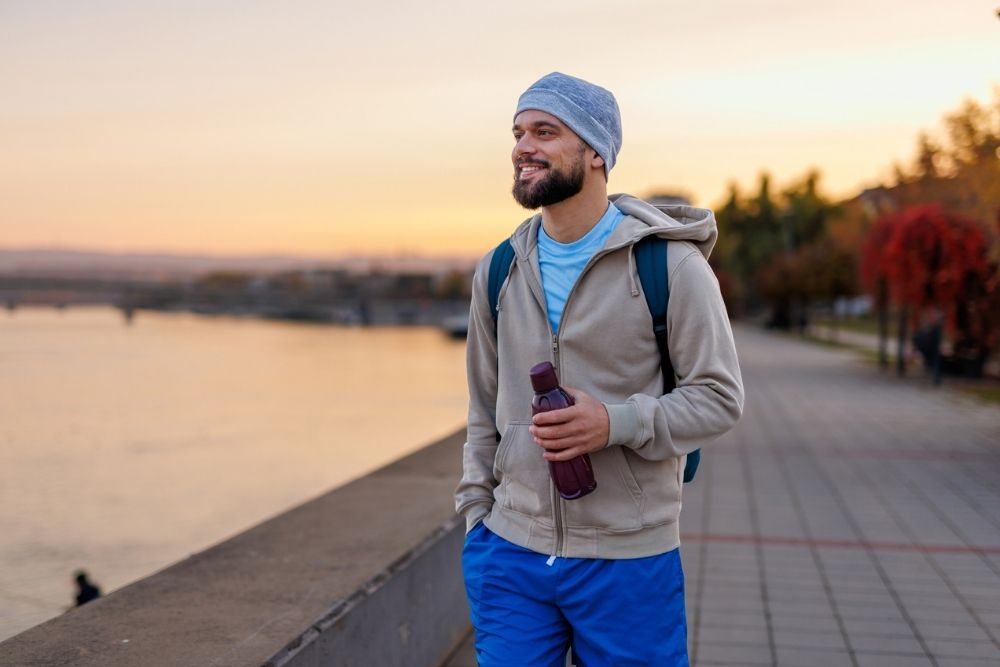 Man walking outdoors at sunset with a water bottle and backpack, smiling and looking relaxed.