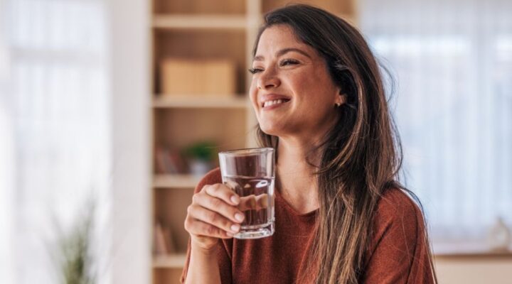 Smiling woman holding a glass of water indoors, enjoying a moment of relaxation and hydration in a bright, cozy room.