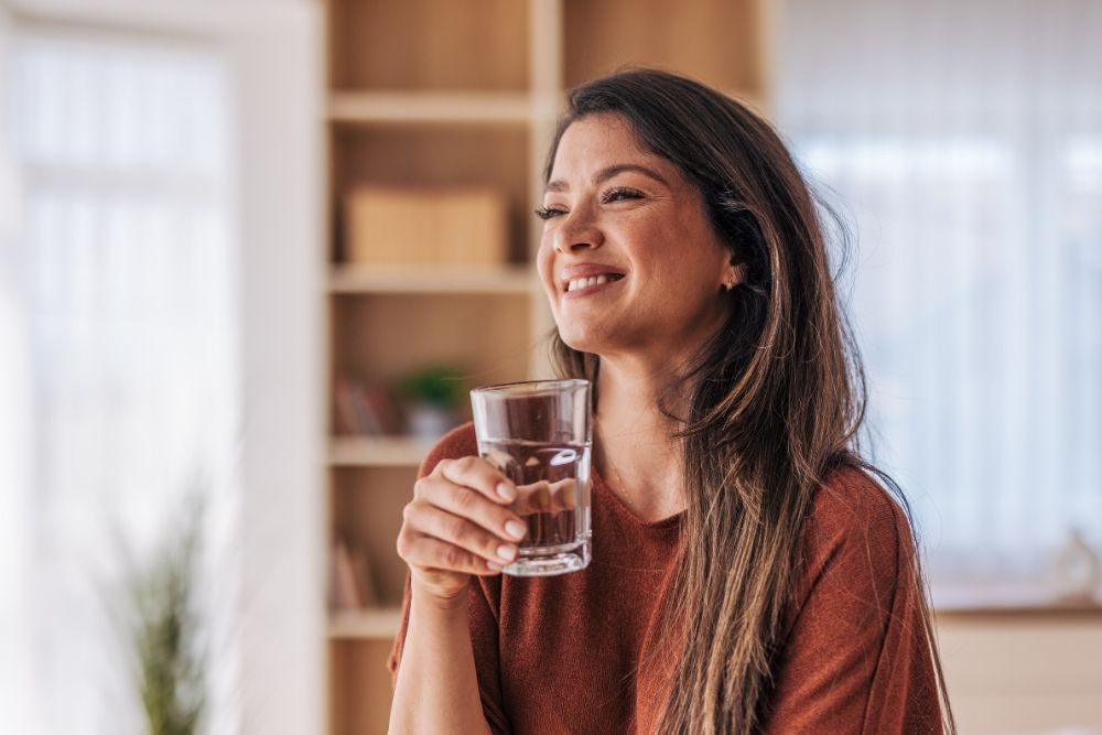Water Hydration(1000 x 667 px) (1) Smiling woman holding a glass of water indoors, enjoying a moment of relaxation and hydration in a bright, cozy room.