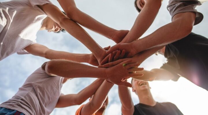 A group of people stand in a circle outdoors, joining hands in the center as a gesture of unity, teamwork, and support in recovery.