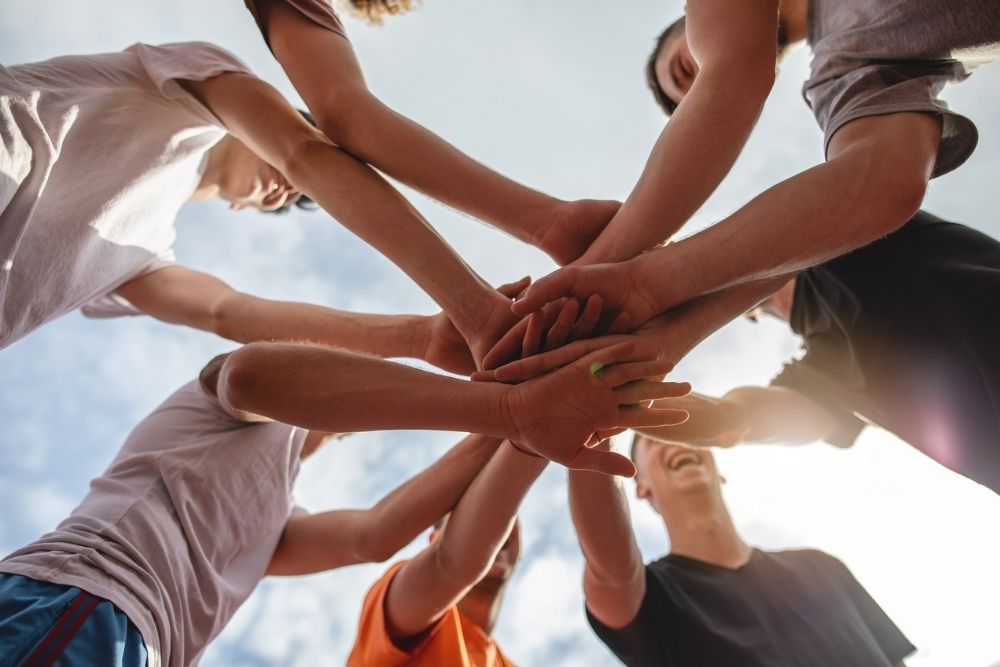 Recovery Competition (1000 x 667 px) A group of people stand in a circle outdoors, joining hands in the center as a gesture of unity, teamwork, and support in recovery.