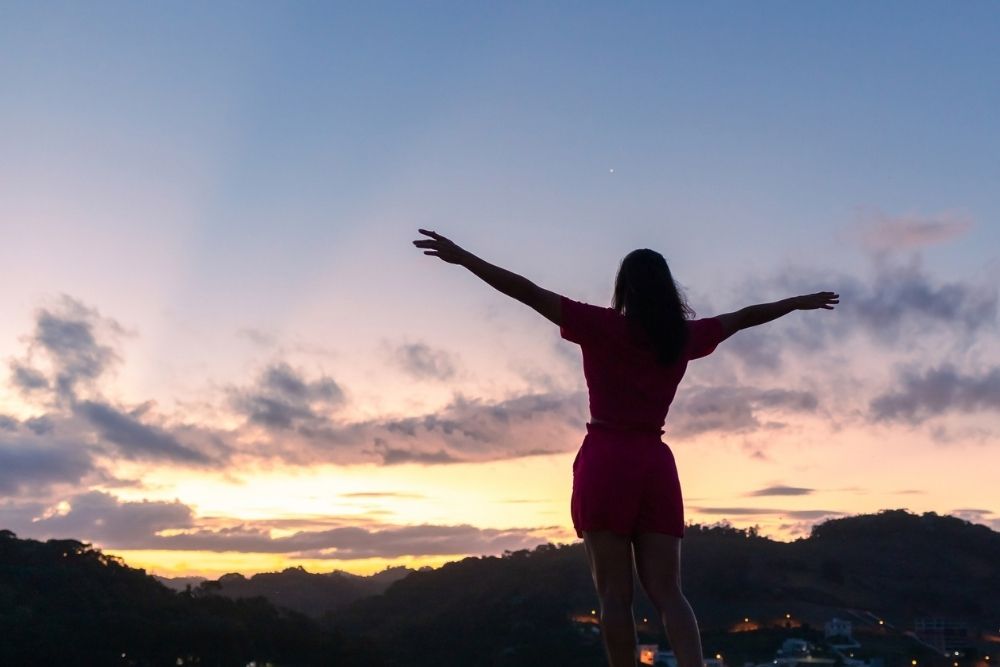 A woman stands outdoors at sunrise or sunset with her arms outstretched, silhouetted against a colorful sky, expressing freedom and resilience.