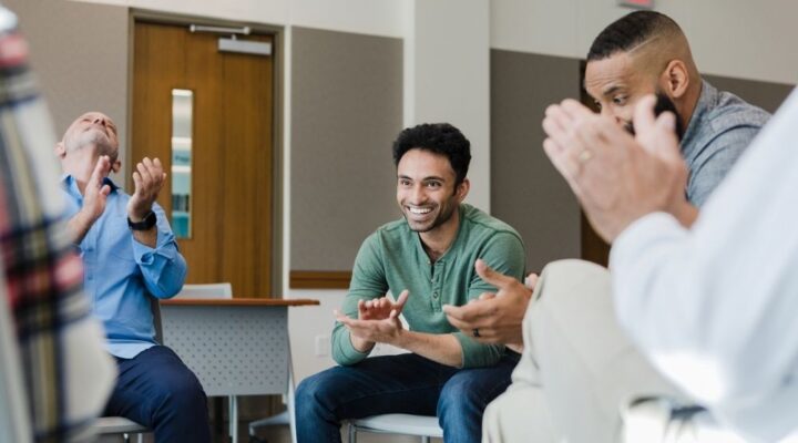 A group of men sitting in a circle, smiling and clapping during a supportive group therapy or recovery meeting.
