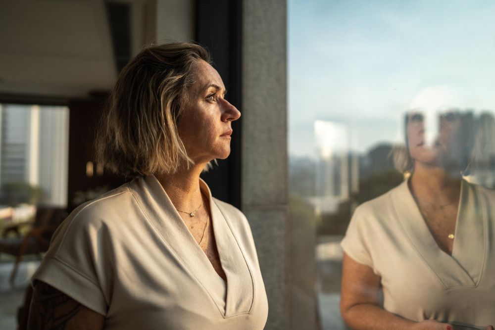 Woman Thinking(1000 x 667 px) A woman stands by a window looking outward with a thoughtful expression, her reflection visible in the glass.