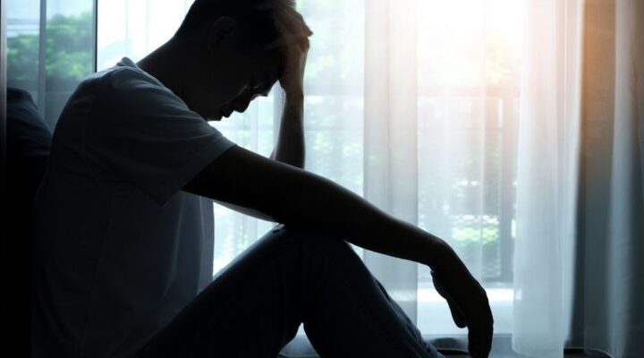 Bel Aire - Mixing Alcohol With Other Drugs Increases the Danger. A man sits in front of a high-rise window with his head in his hand. He is in a shadow position.
