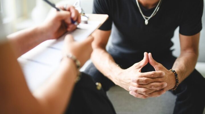 A person takes notes while another sits with interlocked fingers during a conversation.